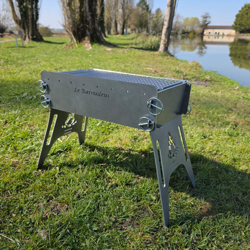 Le barbecue portable Le Baroudeur BBQ monté sur l'herbe avec le logo en façade et un lac en arrière-plan